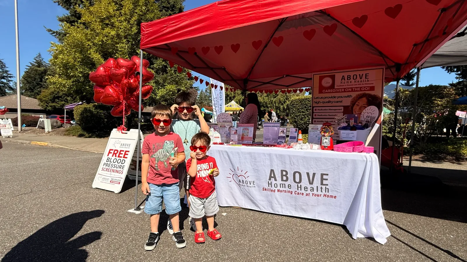 Three children in heart-shaped sunglasses standing in front of the ABOVE Home Health community outreach booth, with a red canopy, heart balloons, and a free blood pressure screenings sign